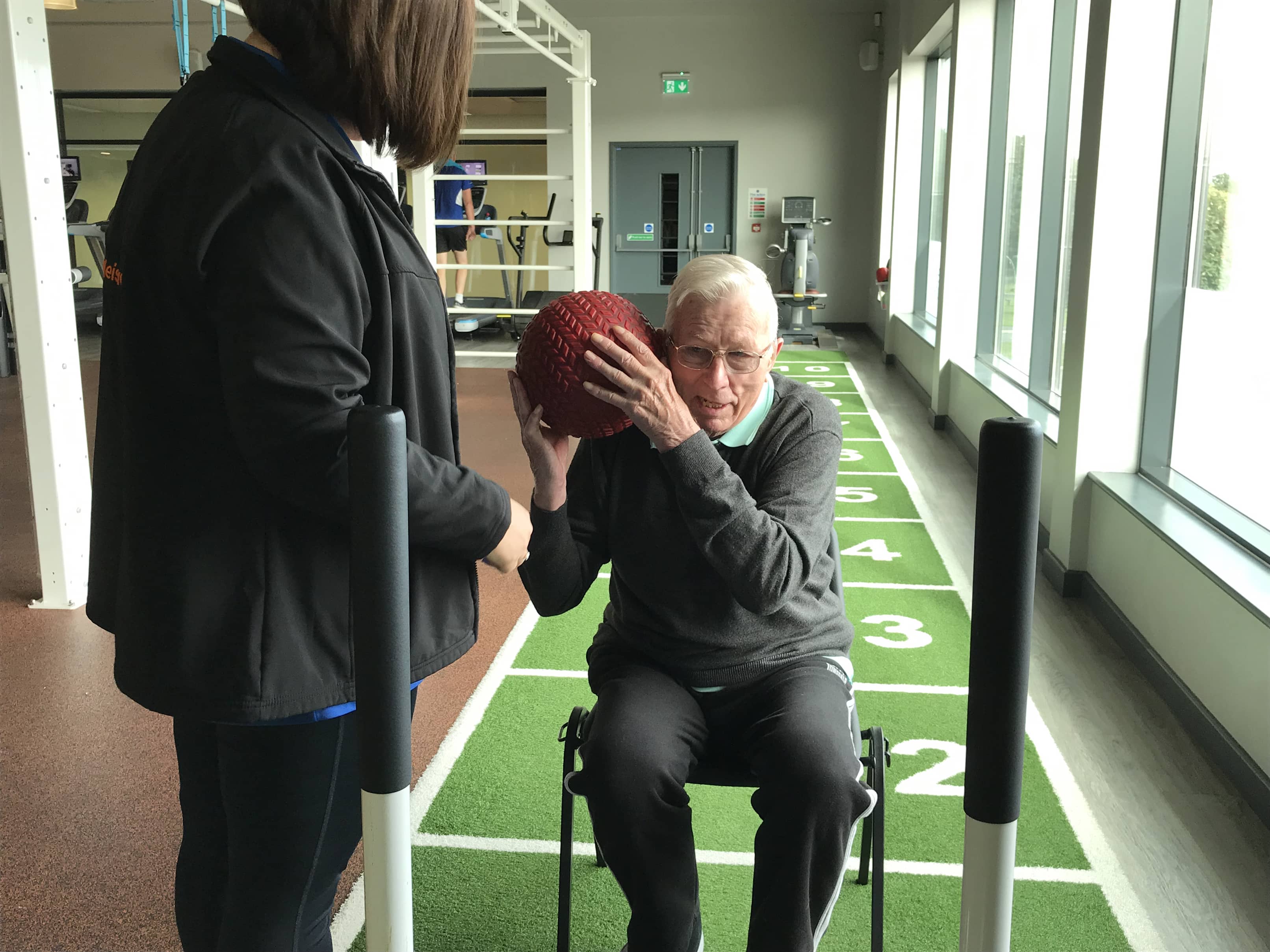 older gentleman throwing slam ball over shoulder whilst sat down accompanied by fitness instructor